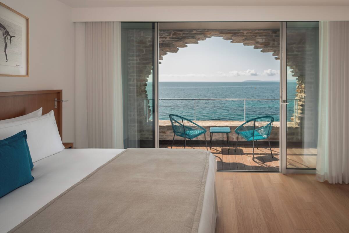 Habitación de hotel con una cama grande, vista al mar a través de un ventanal y dos sillas afuera.