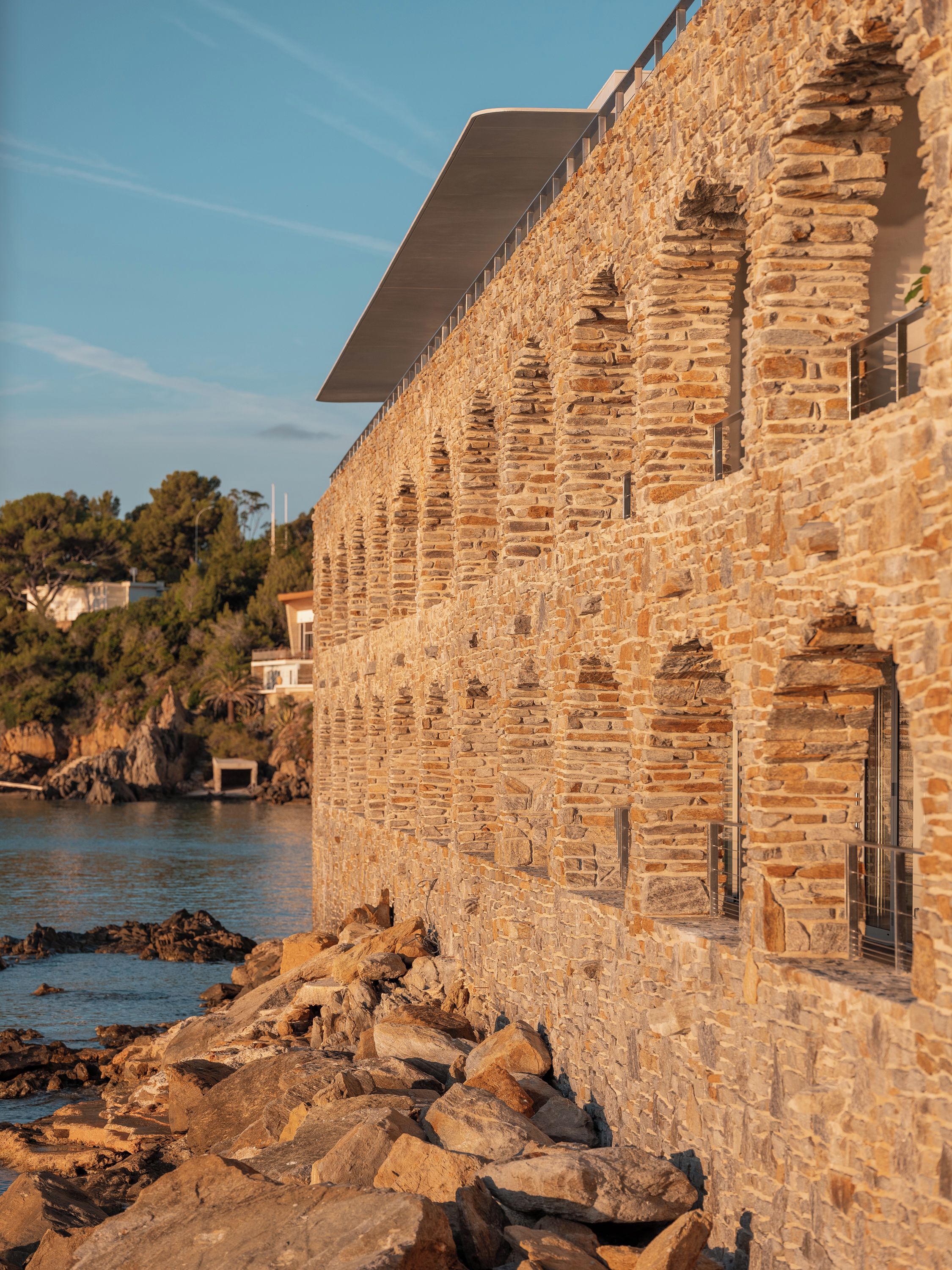 A stone building with arched windows and a flat roof, located near a rocky shoreline.