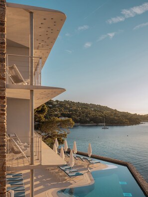 A modern resort with a pool, lounge chairs, and a view of the sea and hills. - Hôtel Les Roches – Relais & Châteaux (Le Lavandou)