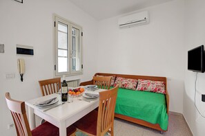 Une pièce comprenant une table à manger dressée pour deux personnes, un lit recouvert d’une couverture verte, un téléviseur fixé au mur et un appareil de climatisation.