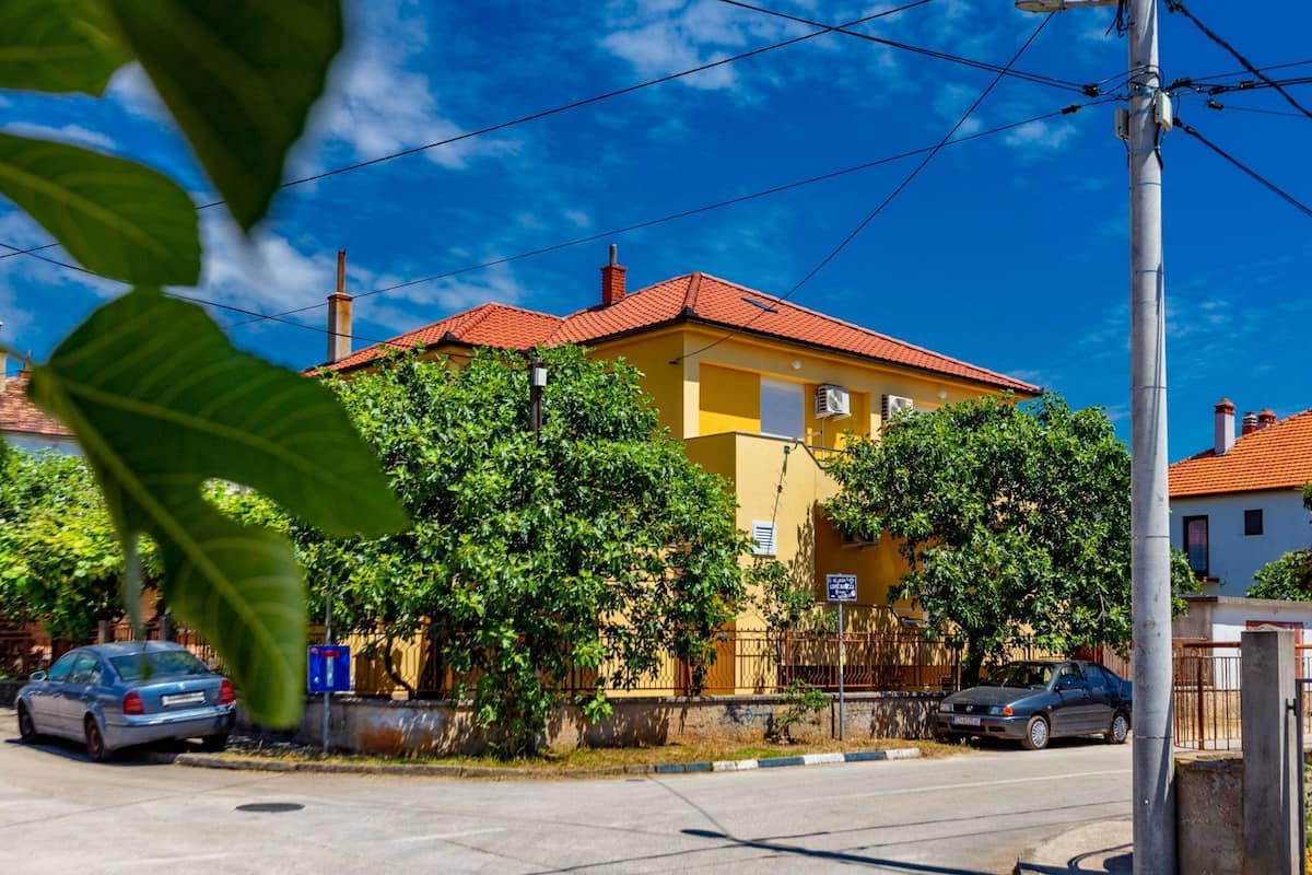 A yellow building with a red roof, surrounded by greenery and parked cars.