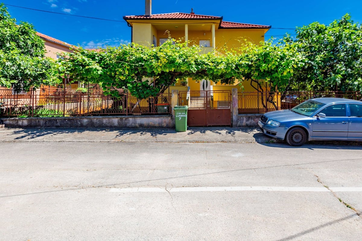 A yellow house with a red roof, a parked car, and a green trash bin.