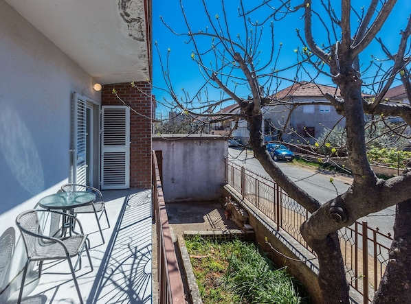A balcony with a table and chairs, a view of a street, and a tree with budding leaves.