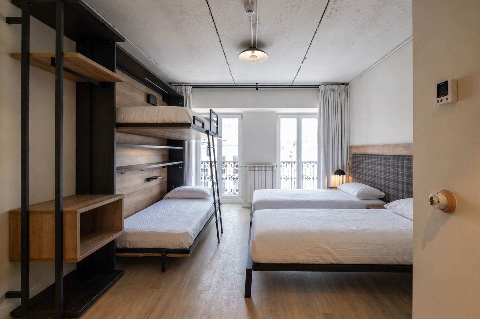A room with bunk beds, a wooden shelf unit, a window with curtains, and a door with a lock.