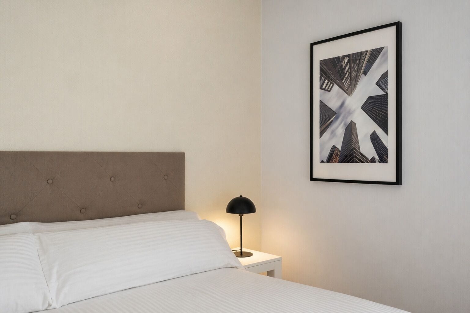 A neatly made bed with a headboard, a bedside lamp, and a framed abstract artwork on the wall.