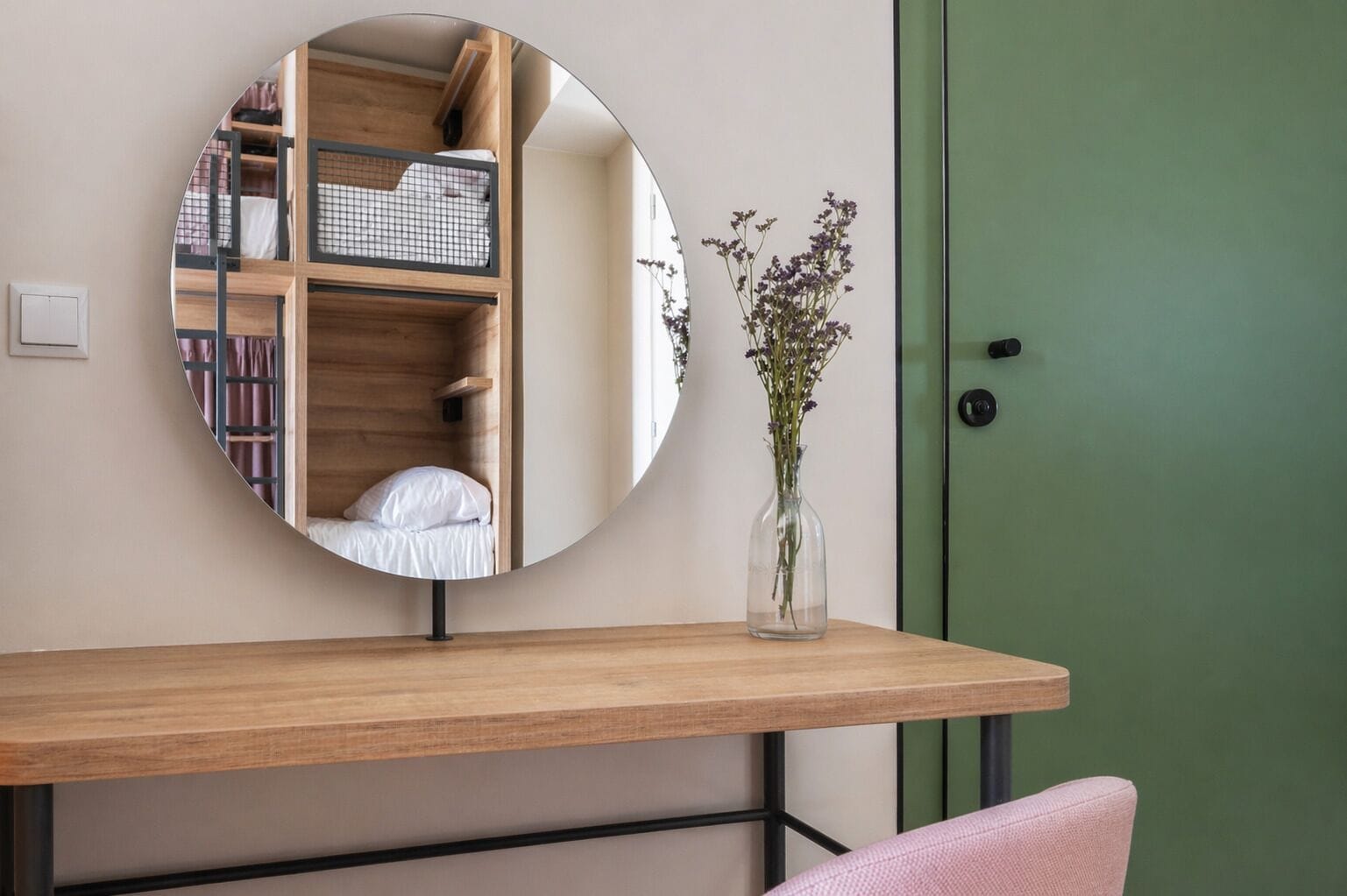 Un miroir rond posé sur une console en bois, agrémenté d’un vase de fleurs, une porte verte et un lit avec un oreiller blanc.
