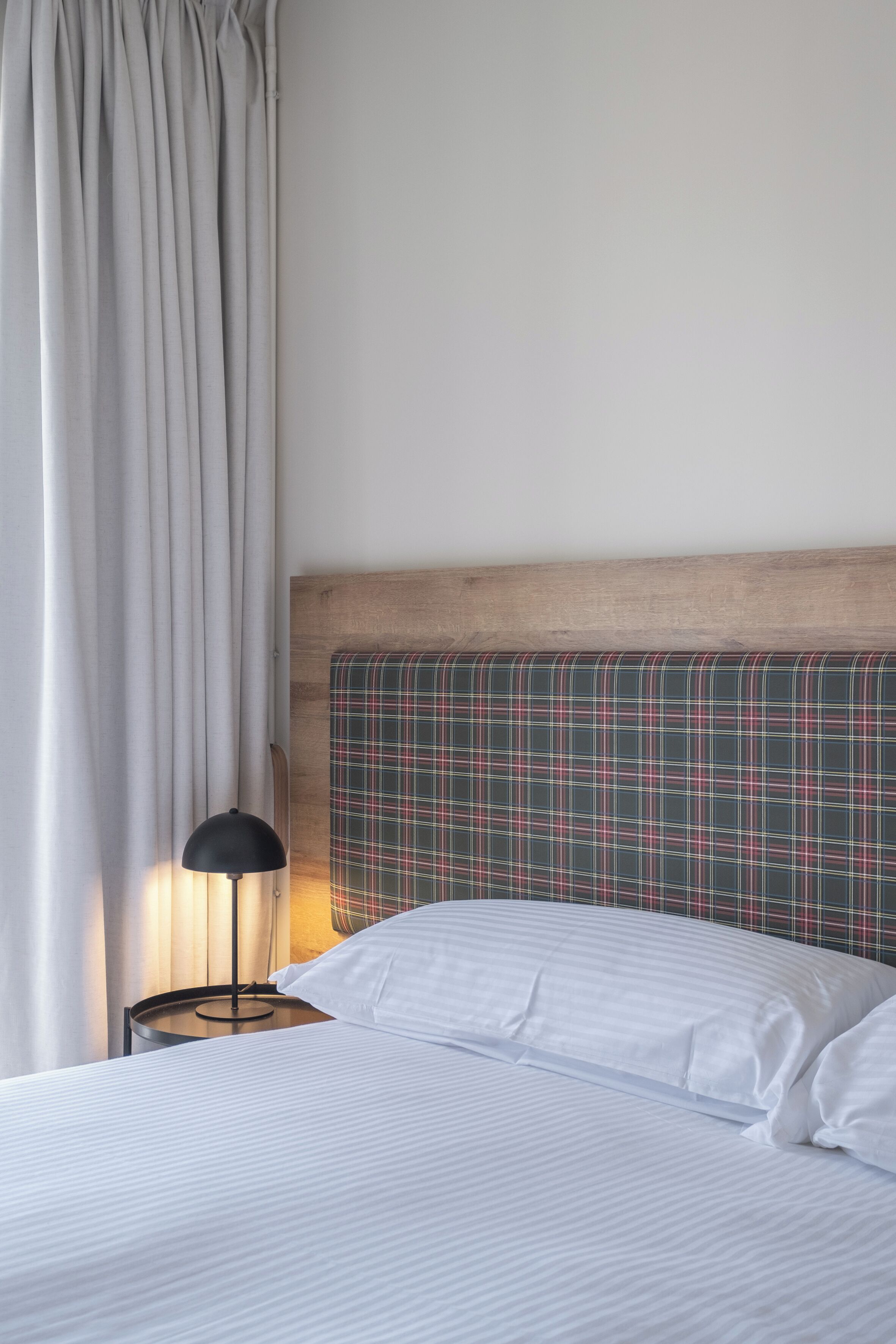 A neatly made bed with white linens, a plaid headboard, a bedside lamp, and a window with curtains.