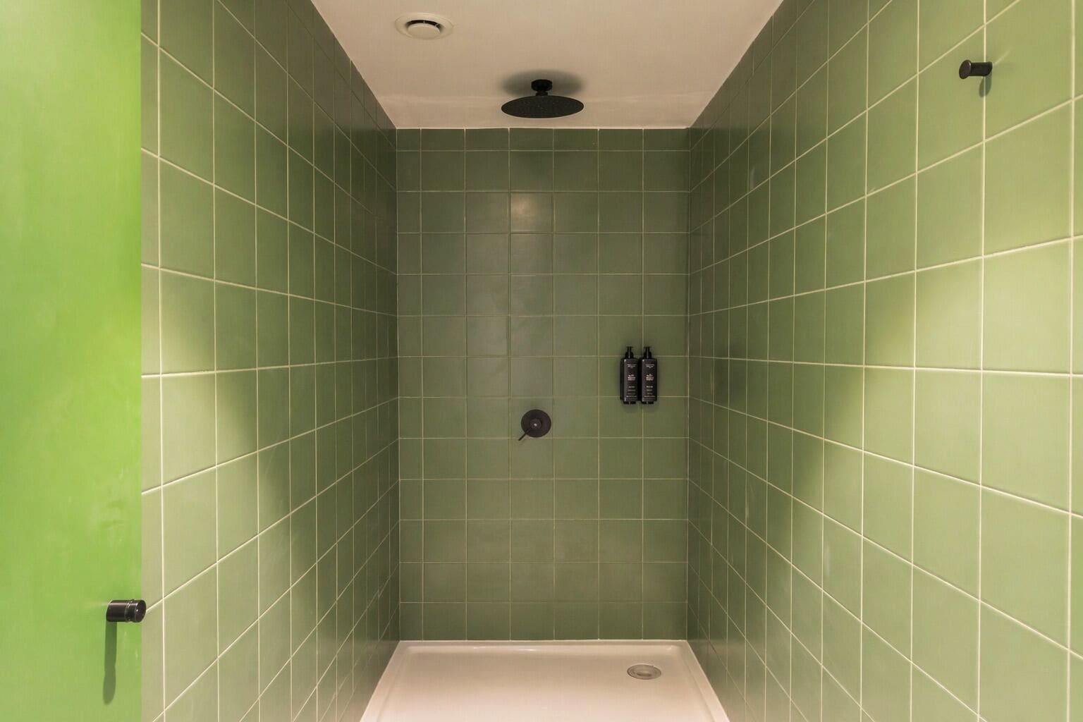 A shower with green tiles, a ceiling-mounted showerhead, and two bottles on the wall.