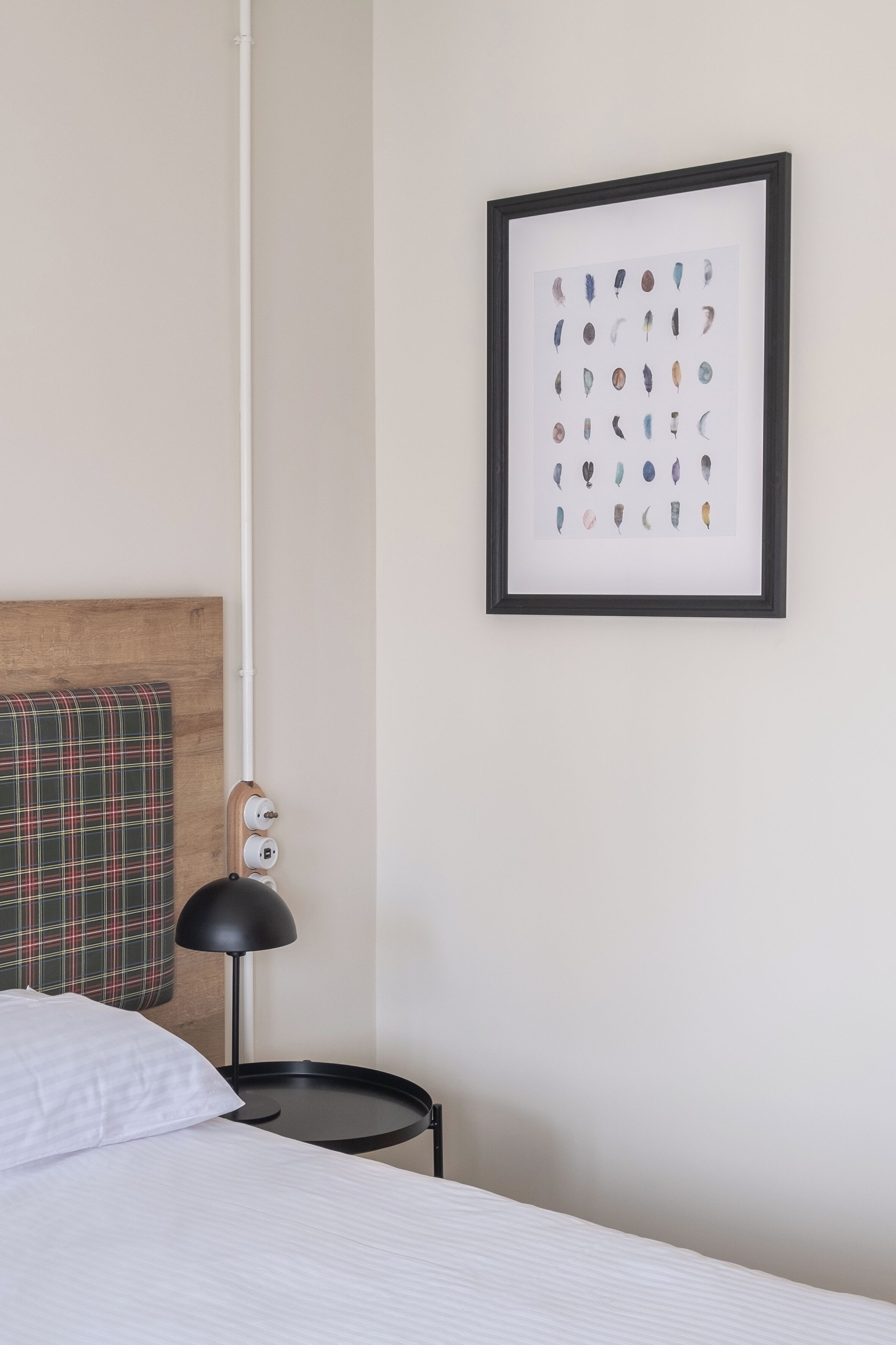 A neatly arranged hotel room with a bed, a framed artwork on the wall, a plaid pillow, and a small bedside table with a lamp.