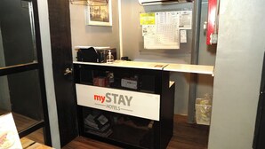 A reception desk with a sign reading "mySTAY HOTELS". There is a framed picture on the wall above the desk, a bulletin board with notices, and a small trash bin to the right. - mySTAY BGC North (Makati)