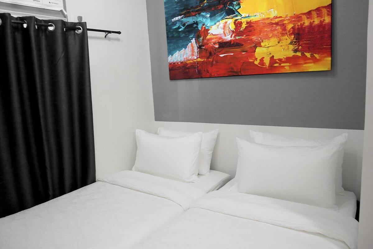 A hotel room with a bed, white bedding, a black curtain, and a colorful abstract painting on the wall.