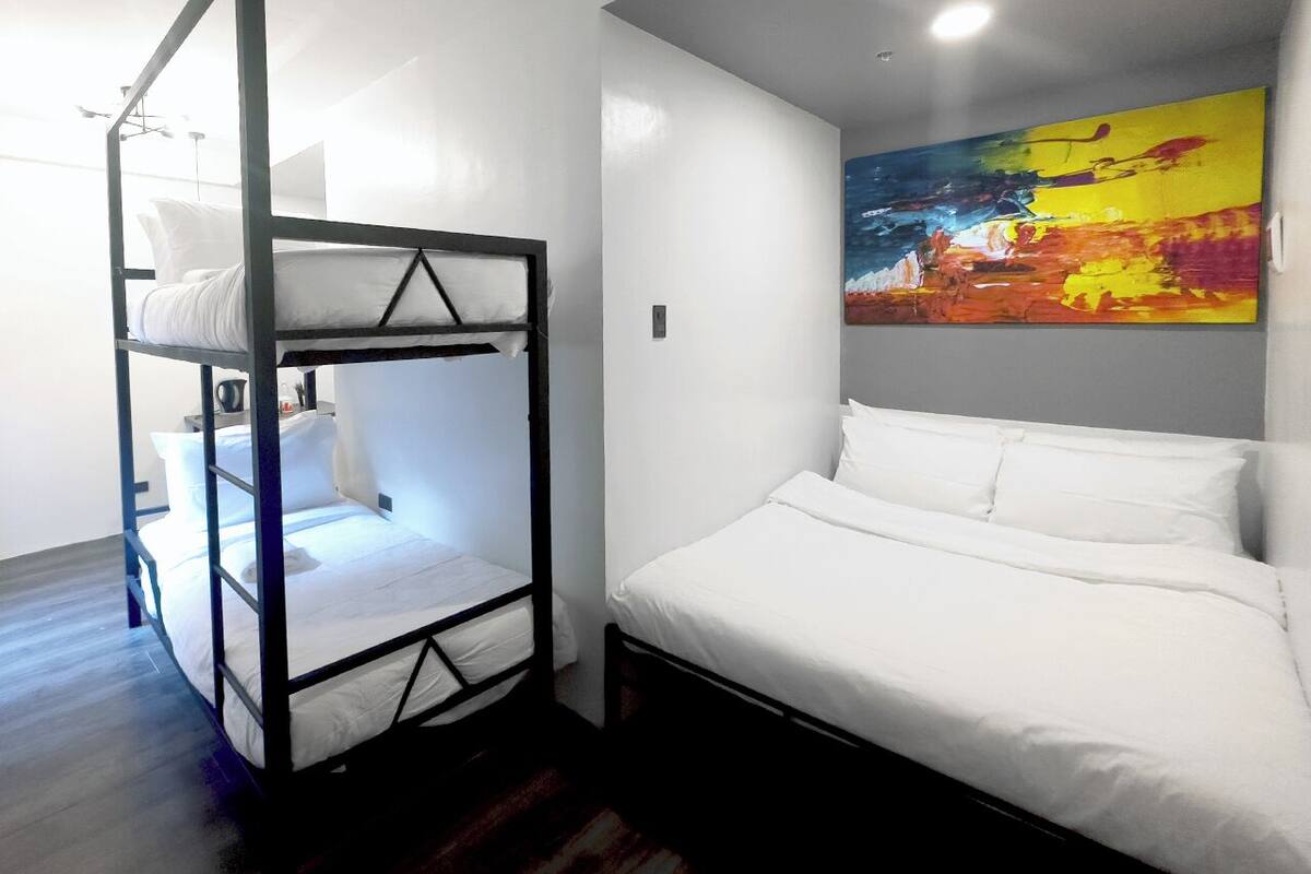 A room with a bunk bed and a single bed, both with white bedding. There is a colorful abstract painting on the wall.