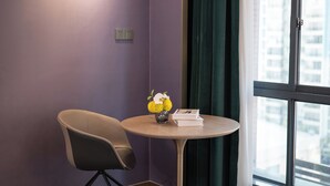 A room with a purple wall, a round table with a vase of flowers, a chair, and a window with a view of buildings. - NanNing Mandala Hotel (Nanning)