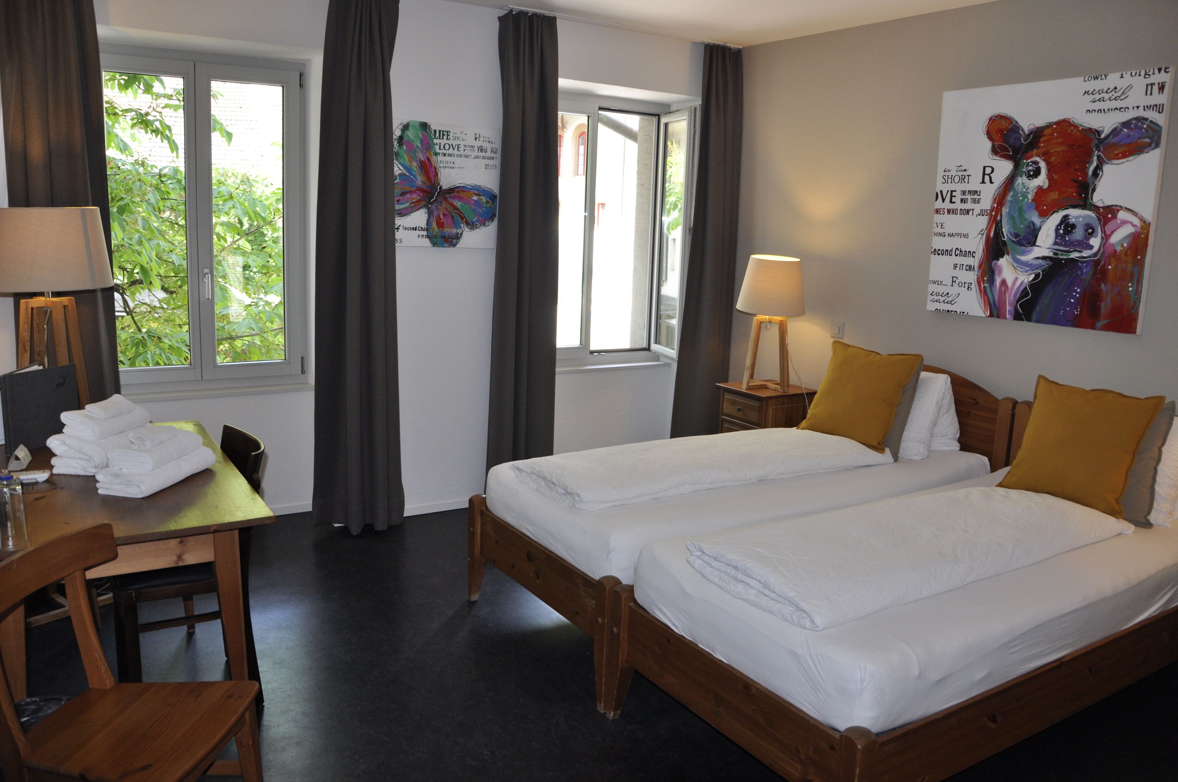 A hotel room with two beds, a desk with a chair, a table with towels, and a window with curtains.