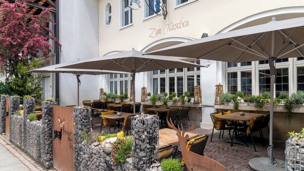Ein Außenbereich mit Tischen und Stühlen, umgeben von einer dekorativen Steinmauer und Kübelpflanzen.