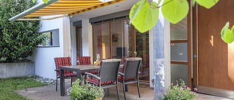 Un patio exterior con mesa y sillas, una planta en maceta y un ventanal de vidrio que da a un espacio interior.