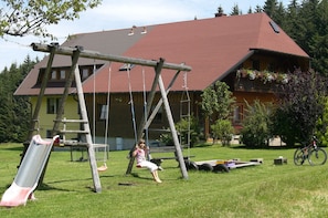Un ensemble de jeux en bois comprenant une balançoire et un toboggan, ainsi qu’un vélo, dans une cour verdoyante, avec une maison en arrière-plan.