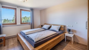 Une chambre à coucher avec un lit en bois, deux fenêtres, une table de chevet et une vue sur les arbres.