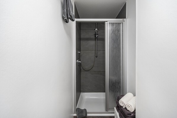 A shower with a handheld showerhead and a wall-mounted faucet, a towel rack with folded towels, and a small towel on the floor.