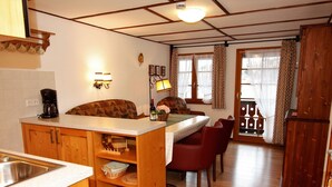 Una cocina con armarios de madera, cafetera, zona de comedor con mesa y sillas, y vista al exterior a través de una ventana.