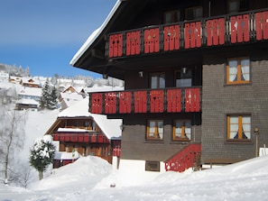 A hotel with a red balcony, snow-covered surroundings, and a view of other buildings in the background.