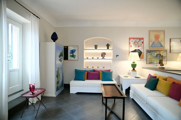 A modern living room with a white sofa, colorful cushions, a small table, and a shelf with decorative items.