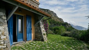 A stone building with blue shutters, a stone staircase, and a garden with a patterned lawn leading to a mountain view.