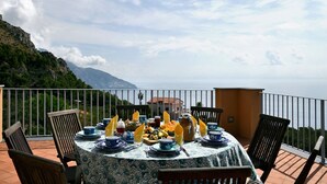 A round table set for a meal with blue and white tableware, surrounded by chairs, on a balcony with a view of the sea and mountains.
