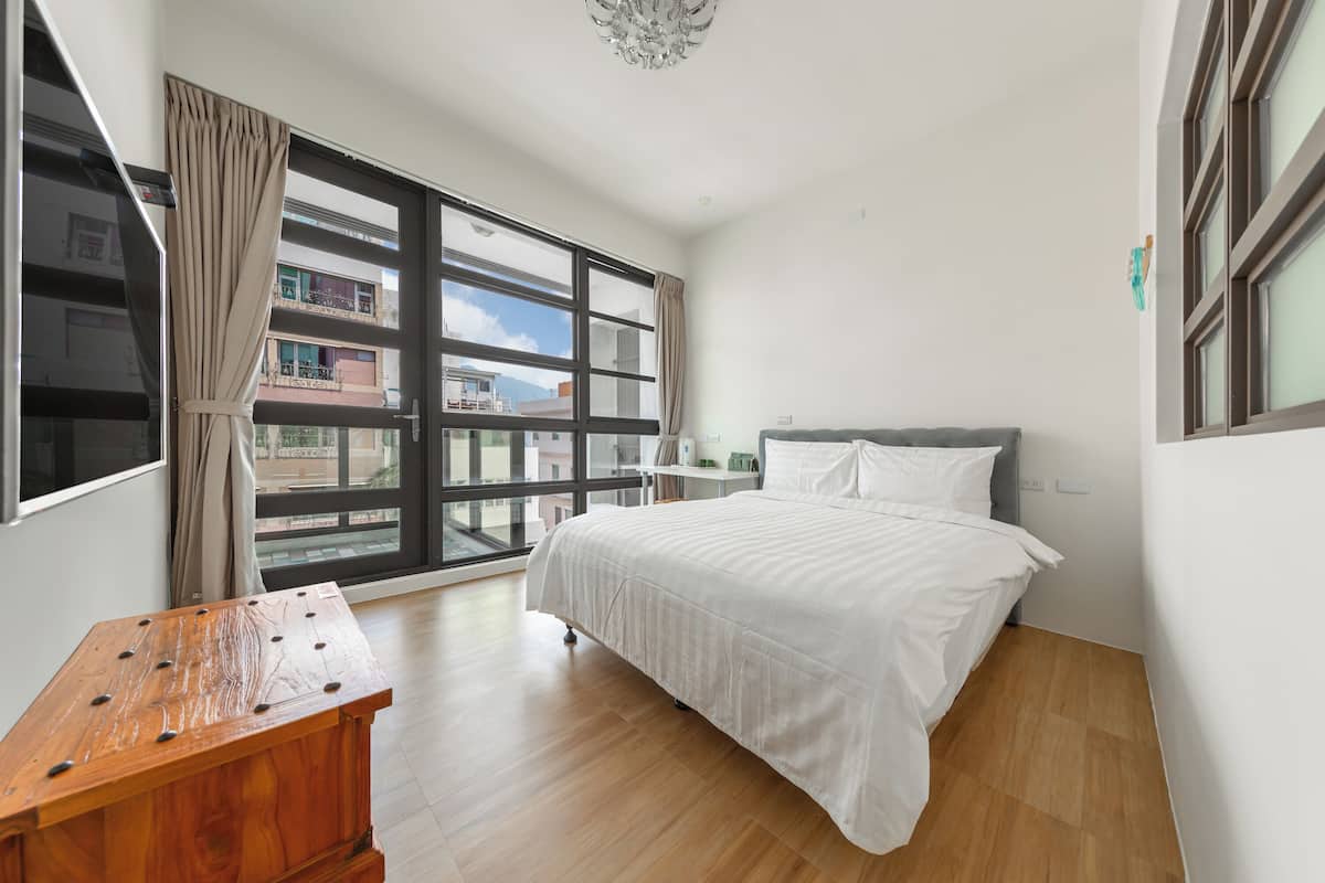 A modern bedroom with a large window, a wooden desk, and a neatly made bed.