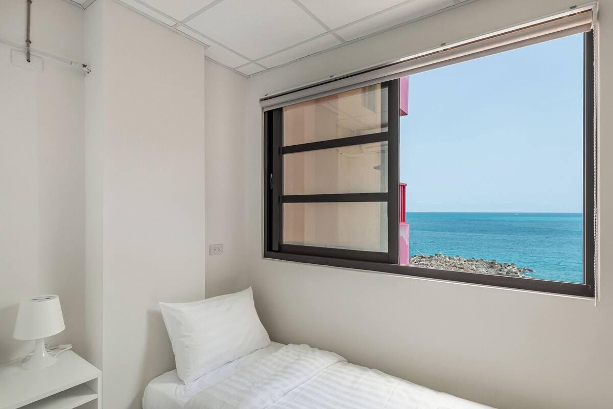 A bedroom with a bed, a nightstand, and a window with a view of the sea.