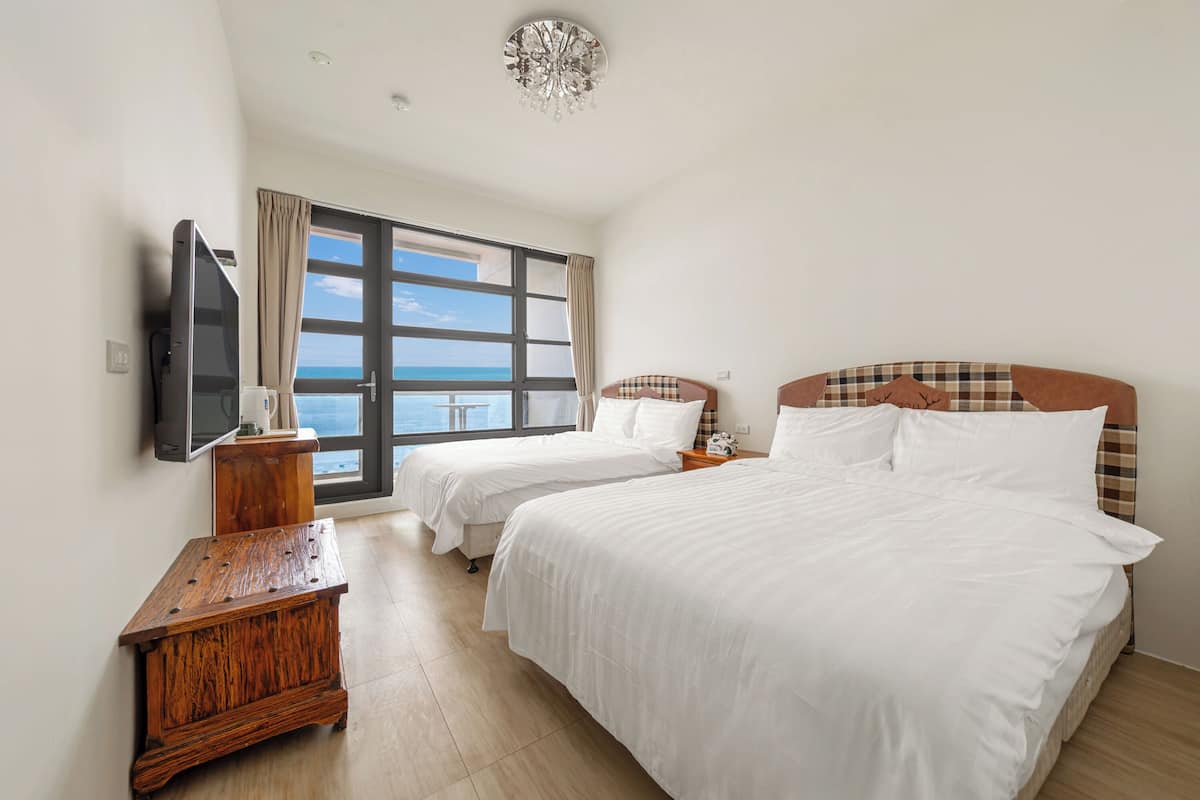 A hotel room with two beds, a TV, a wooden nightstand, and a large window offering a view of the sea.