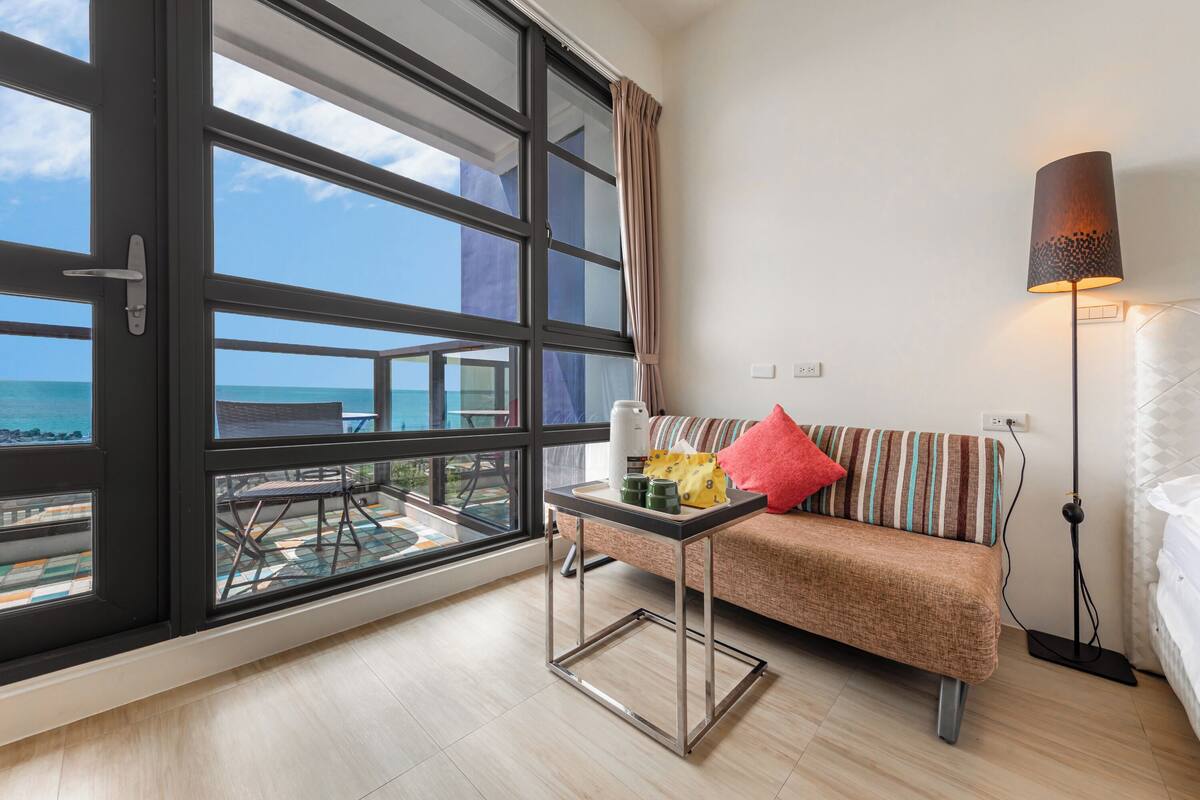 A modern hotel room with a large balcony, a sofa with striped cushions, a small table with a tray, and a floor lamp.