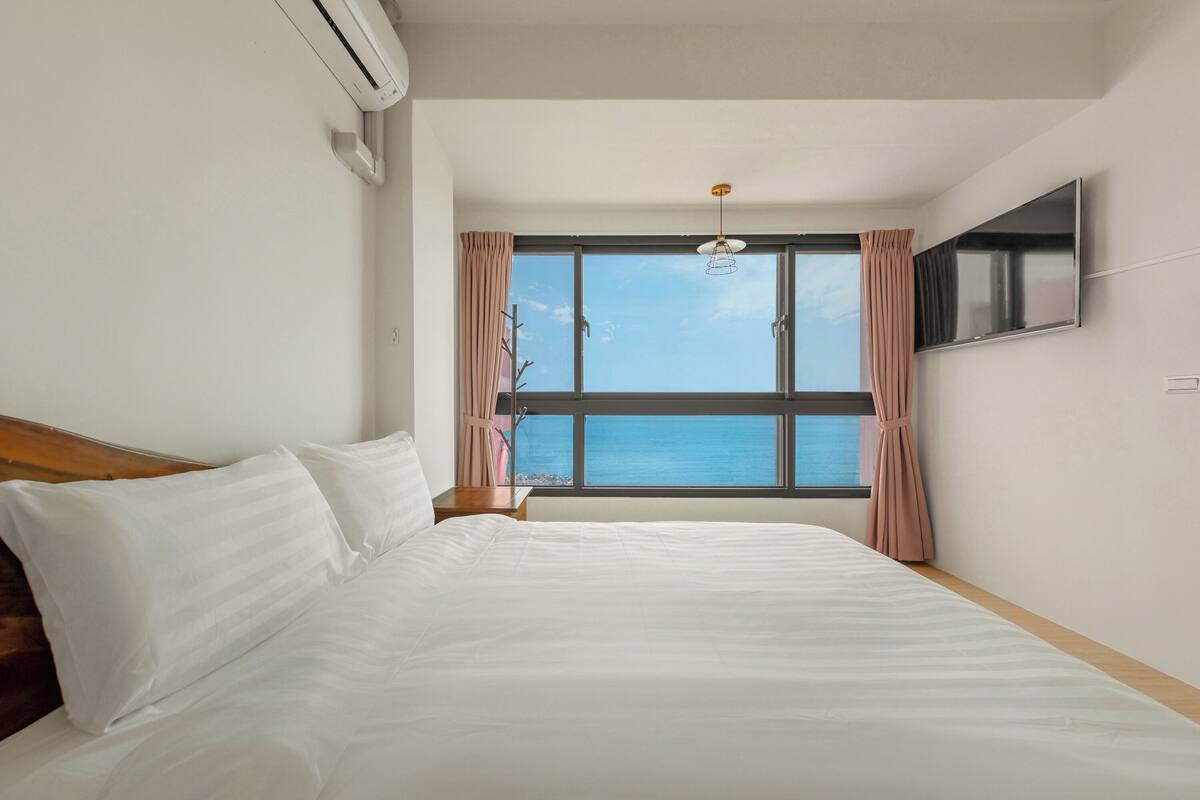 A hotel room with a large bed, a TV mounted on the wall, and a window offering a view of the sea.