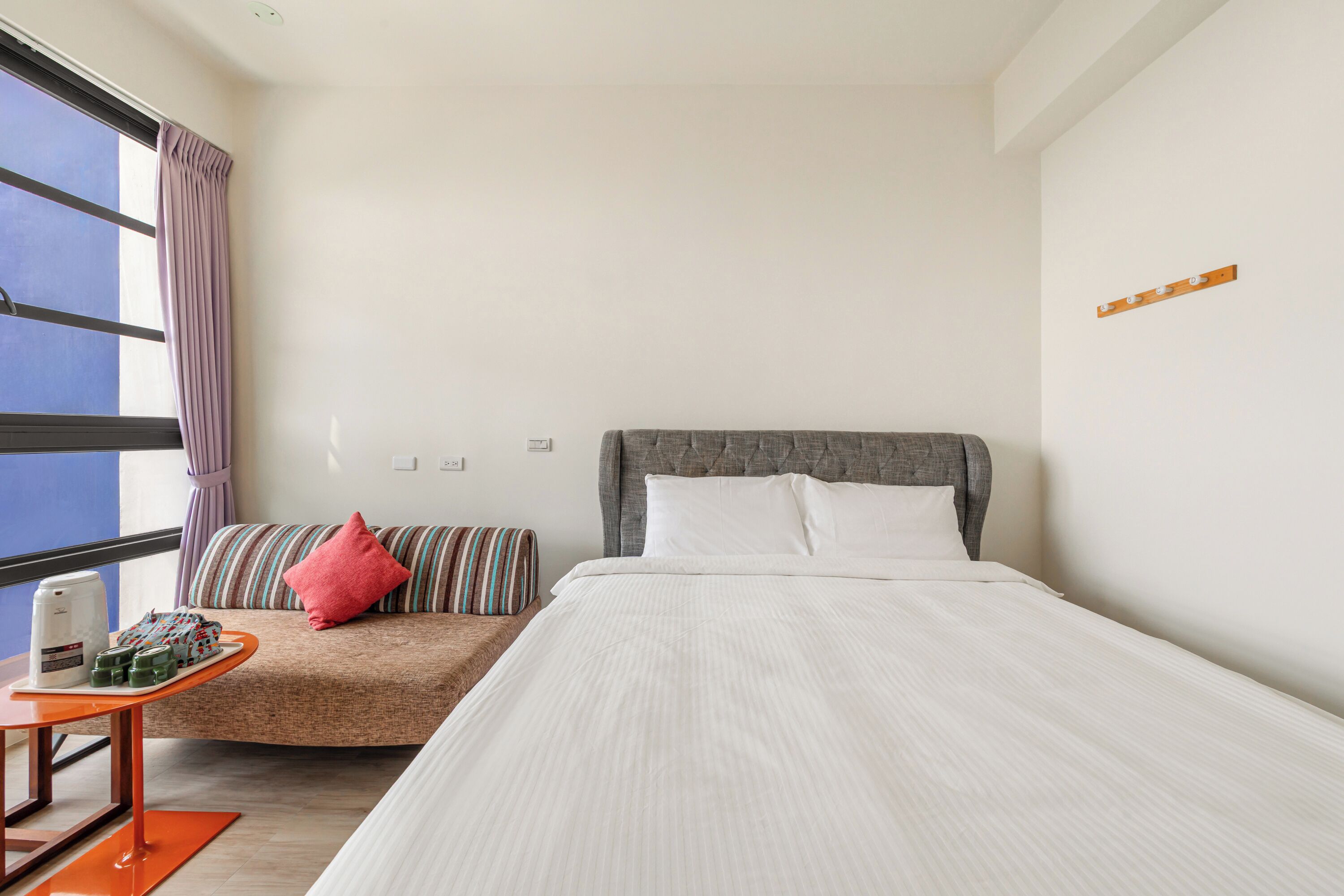 A hotel room with a large bed, a smaller sofa, a table with a coffee maker, and a window with curtains.