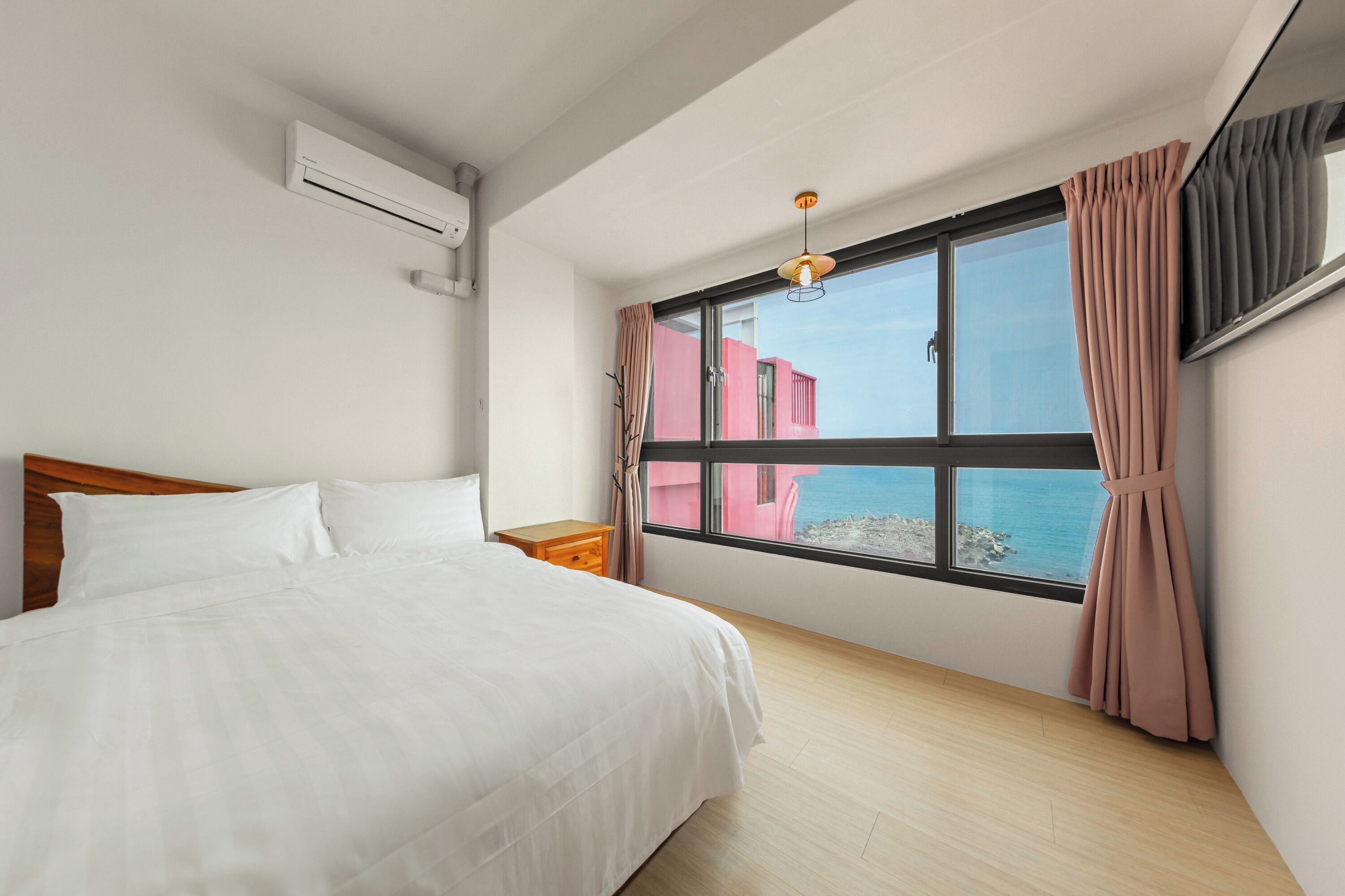 A bedroom with a large bed, a nightstand, a window with curtains, and an air conditioning unit.