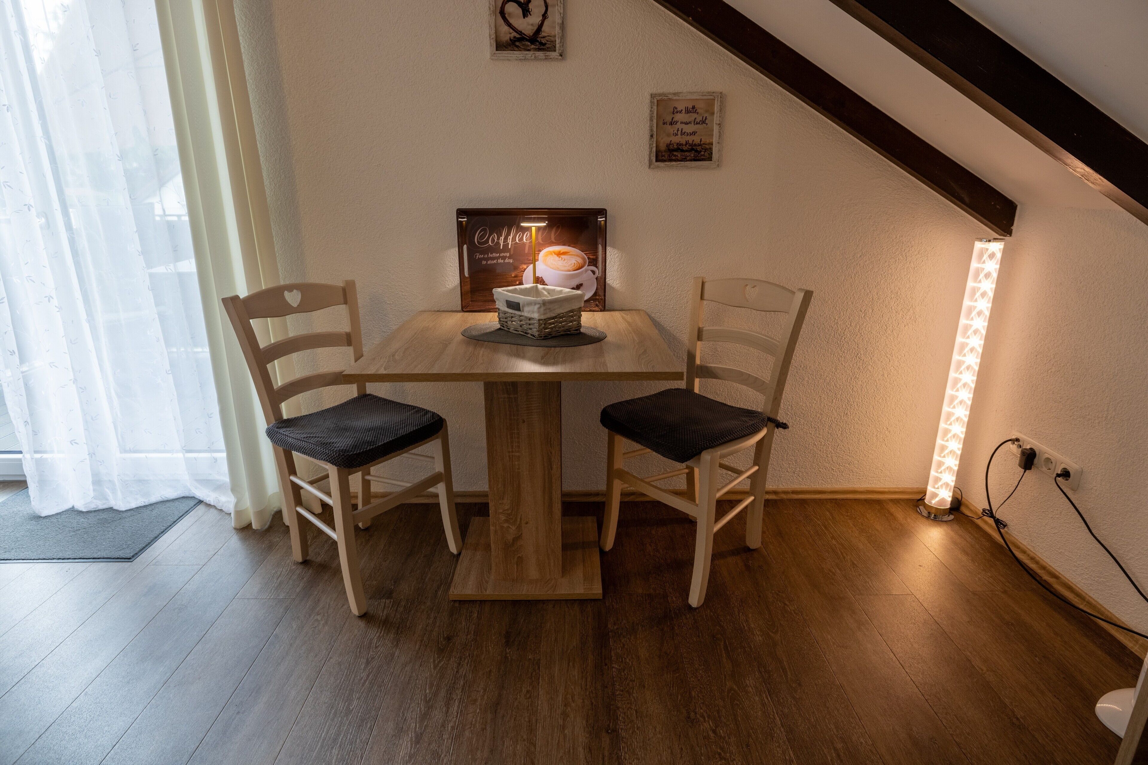 Un coin repas accueillant avec une table en bois et deux chaises, une œuvre d’art encadrée sur le thème du café, et un luminaire fixé au mur.