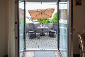 Een glazen deur die uitkomt op een terras met een tafel, twee stoelen en een grote parasol.