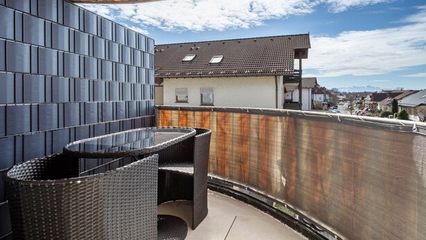 A balcony with a black wicker table and chairs, a metal fence, and a view of a town with houses and mountains in the distance. - Apartment 'Bodenseenest' with Mountain View, Balcony and Wi-Fi (Meckenbeuren)