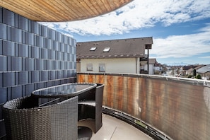 Un balcón con una mesa y sillas de mimbre negro, una valla de metal y vistas a un pueblo con casas y montañas al fondo.