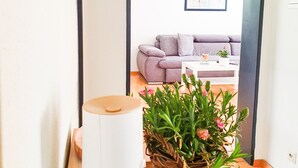 Una habitación con suelo de madera, un objeto cilíndrico blanco, una cesta de mimbre con plantas verdes y un sofá púrpura al fondo.
