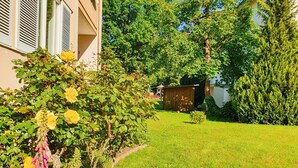 Un jardín residencial con césped, plantas con flores y un edificio con persianas.
