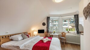 A hotel room with a wooden bed, a red blanket, a small table with a lamp, a chair, and a view of buildings through a window. - Apartment 'Studio' with Wi-Fi (Schluchsee)