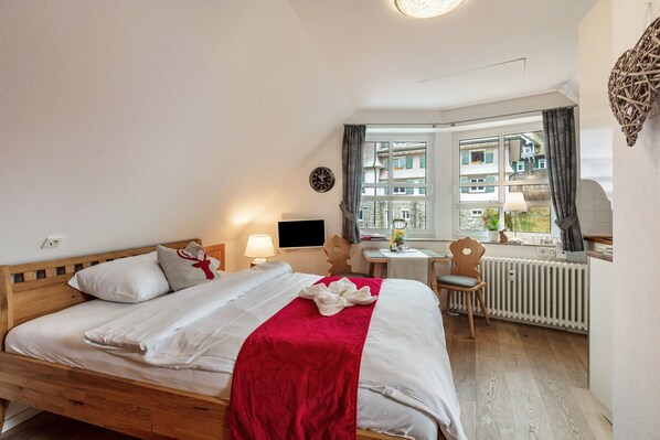 A hotel room with a wooden bed, a red blanket, a small table with a lamp, a chair, and a view of buildings through a window.