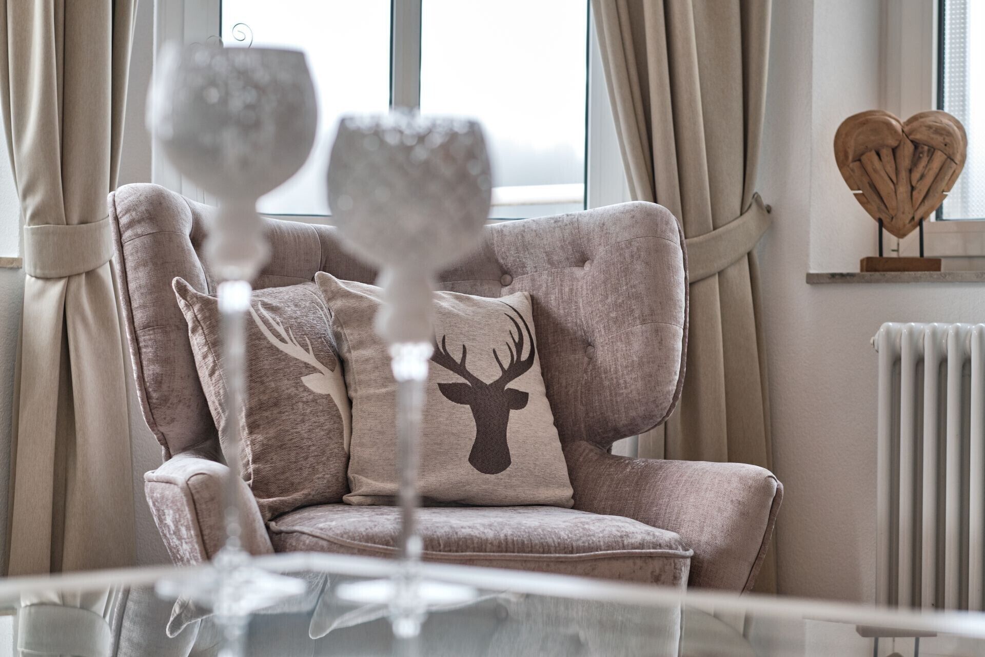 A cozy armchair with a deer pillow, a glass of wine, and a heart-shaped wooden decoration on a shelf.