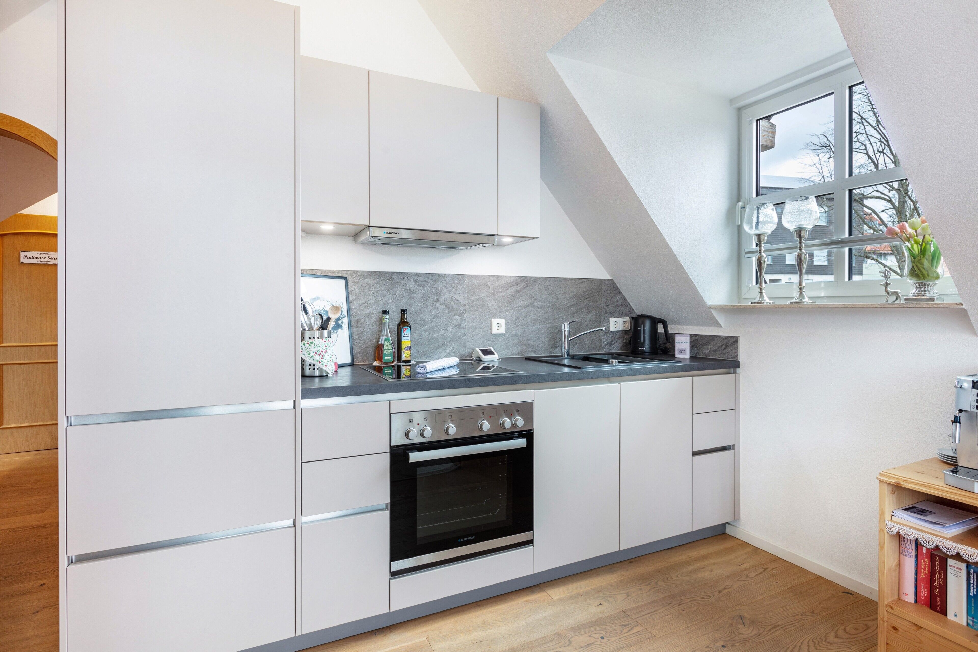 Een moderne keuken met witte kastjes, een ingebouwde oven, een raam met uitzicht en een klein boekenrek.