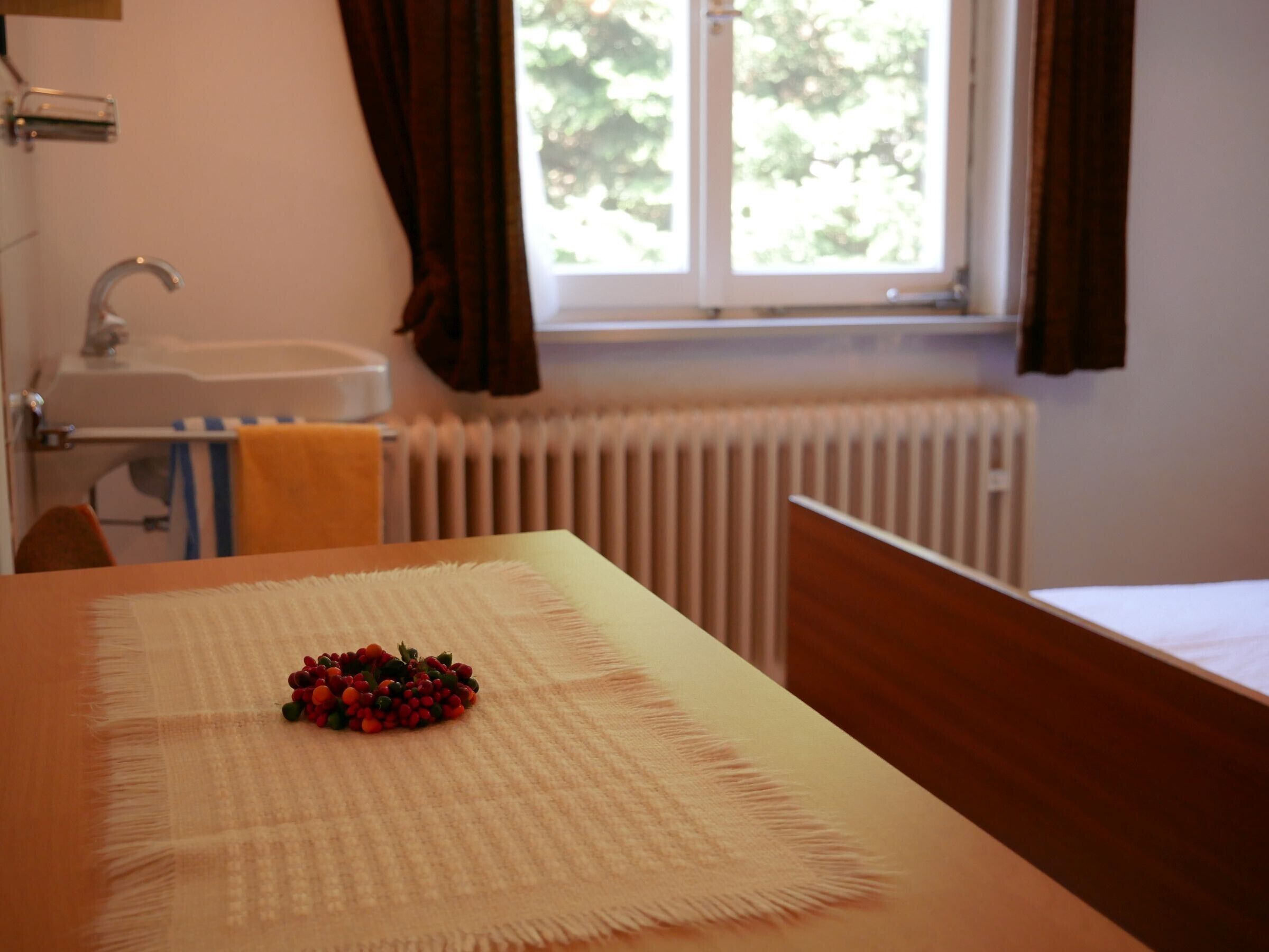 Une chambre d’hôtel avec un lit, une fenêtre avec des rideaux, un radiateur et une table avec un set de table et des fruits rouges.