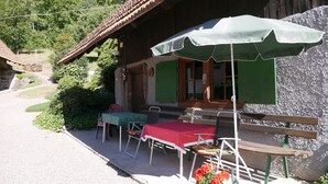A rustic wooden building with a covered outdoor seating area, a table with a red tablecloth, and a potted plant with flowers.