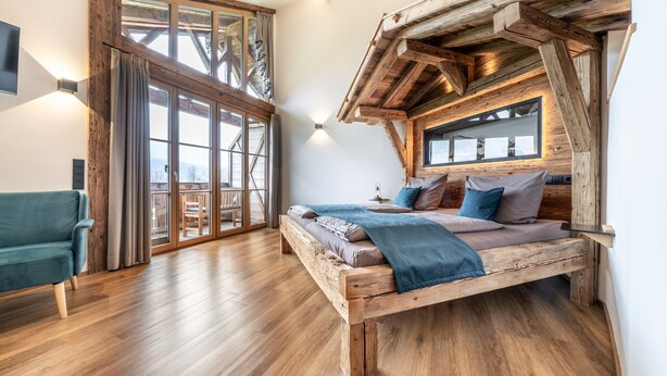 A wooden-framed bedroom with a large window, a bed with pillows, and a blue chair. - Chalet 'Hochsitz' incl. Königscard (Gaißach)