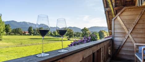 Two wine glasses on a wooden balcony with a view of green fields and mountains.