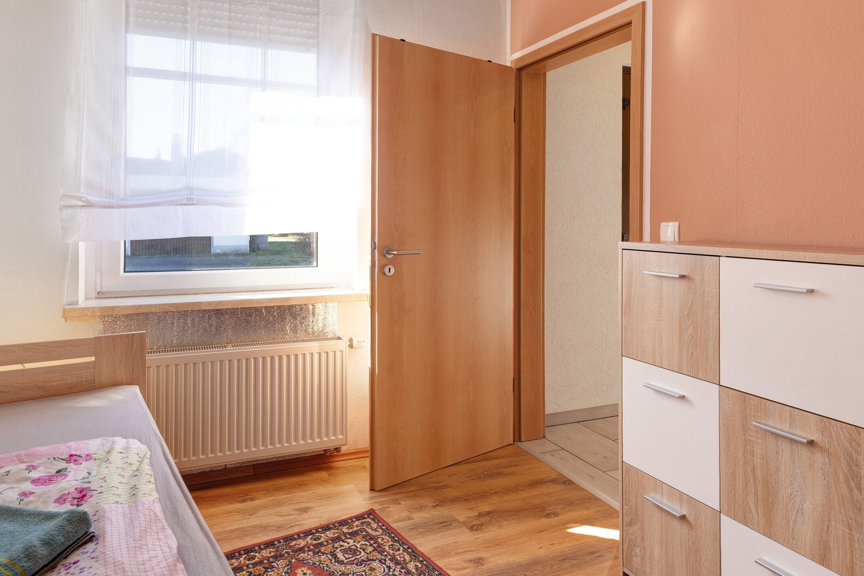 A bedroom with a wooden door, a radiator, a window with sheer curtains, a bed with a floral bedspread, and a wardrobe with multiple drawers.